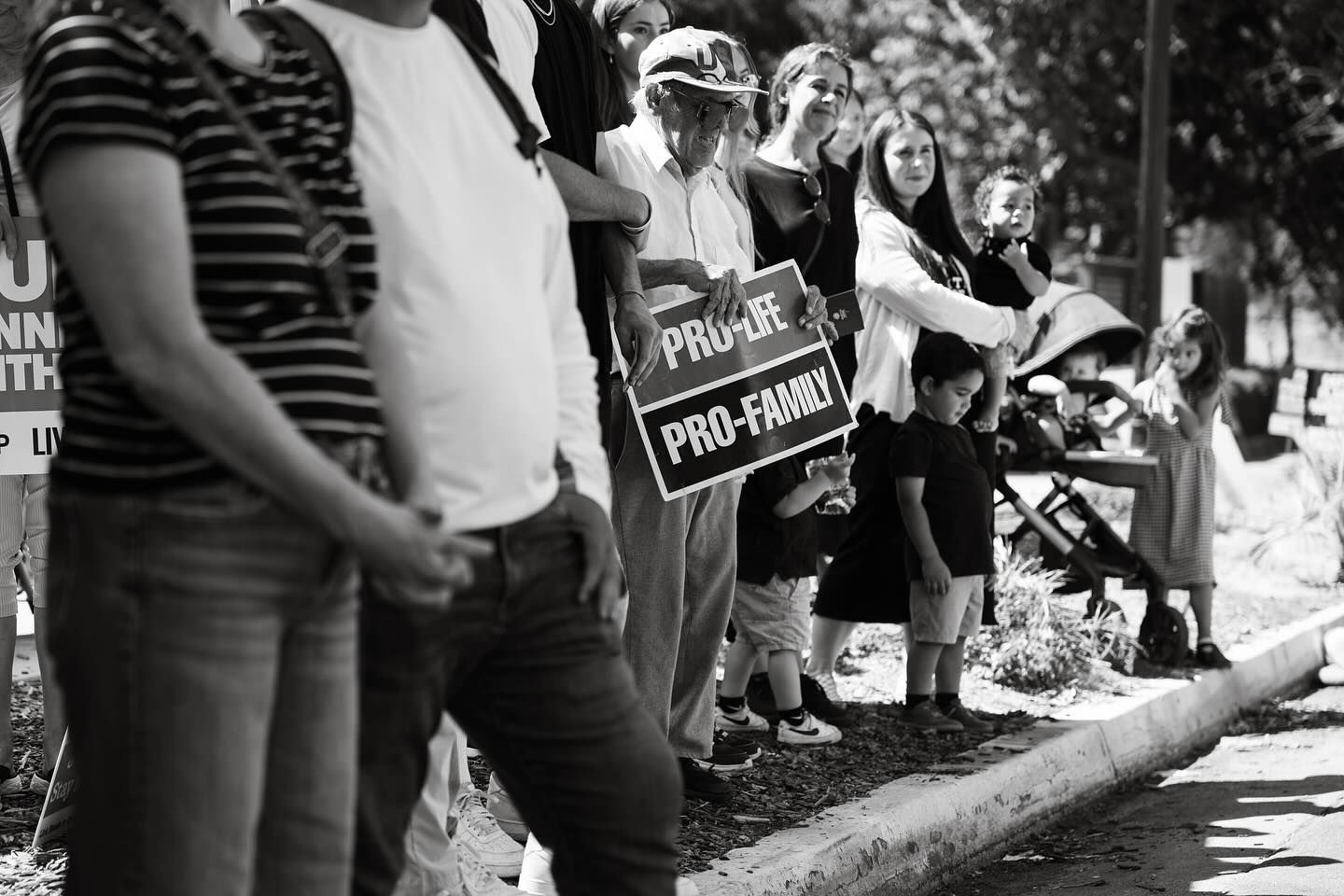 A line of supporters along a curb: an older man holding a Pro-Life, Pro-Family sign, mothers, and small children beside a stroller.
